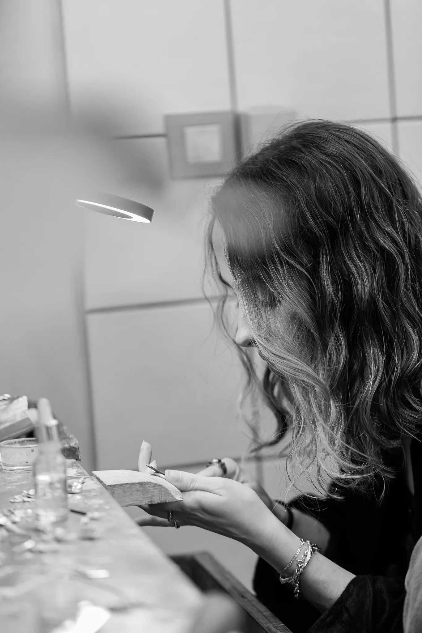Sara concentrada à bancada, iluminada pelo candeeiro de trabalho.
